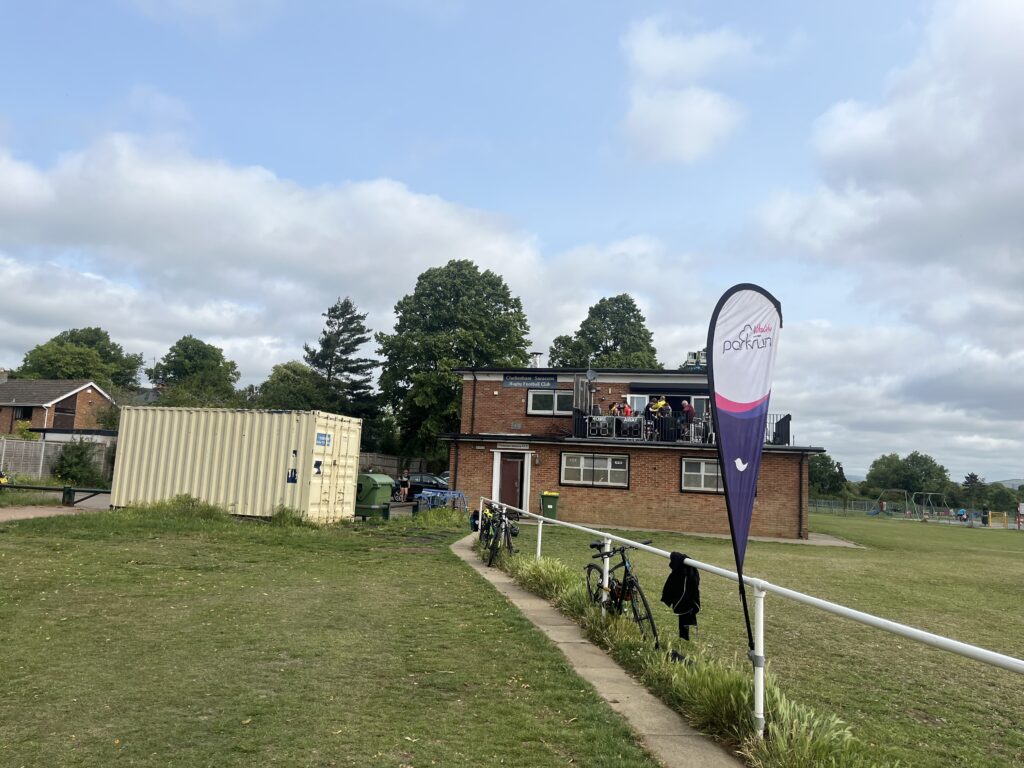 King George V Playing Field parkrun, Parkrun, Running, Runners, A green field with a flag saying parkrun in the foreground with a two storey brick building in the background and people just visible on the roof terrace