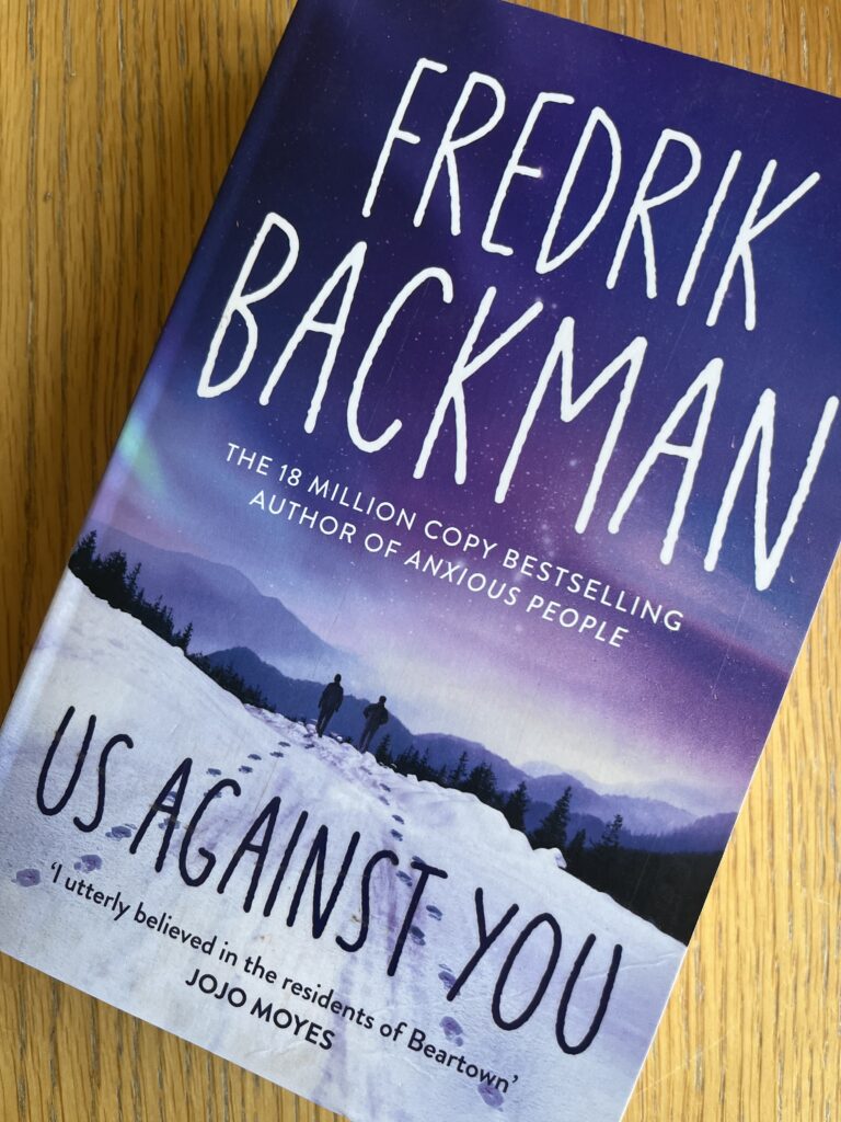 Us Against You by Fredrik Backman, Us Against You, Fredrik Backman, Book review, A blue and purple book with snow on the cover called Us Against You by Fredrik Backman sits on a wooden table