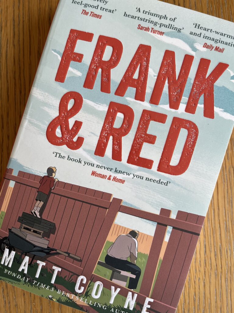 Frank & Red by Matt Coyne, Frank & Red, Matt Coyne, Book review, A book called Frank & Red by Matt Coyne showing a little boy standing on a wheelbarrow looking over the fence at a man sat down looking away from him sits on a wooden table