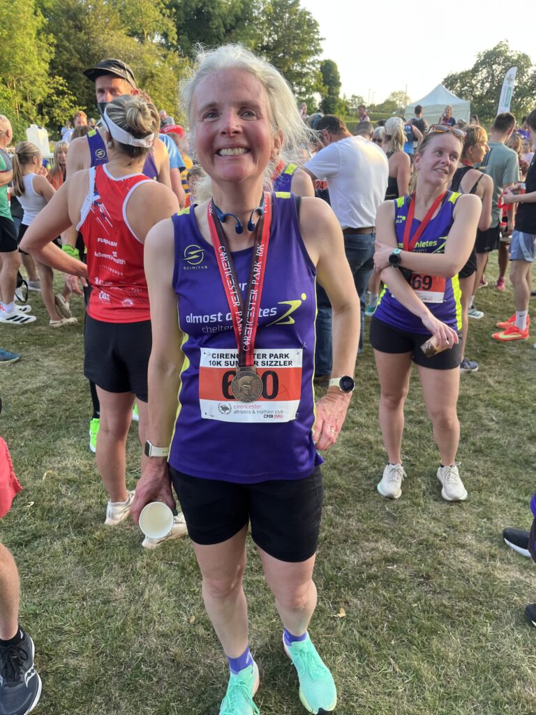 Cirencester Summer Sizzler, Cirencester 10k, Running, Runner, A smiling female runner in a purple vest with a medal round her neck stands on grass in front of crowds of runners