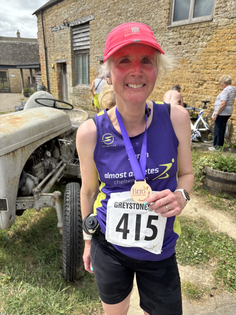 Greystones 10k, Medal, Runner, A female runner in a purple vest and pink hat wearing a wooden medal stands in front of an old fashioned tractor and a Cotswold stone farm building
