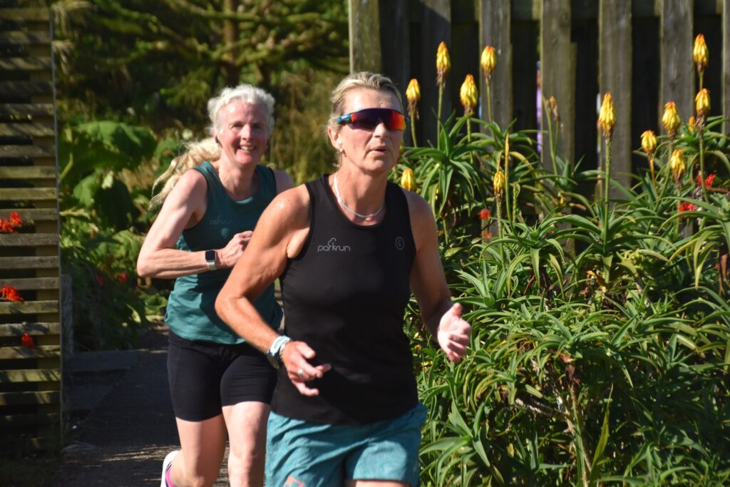 Heartlands parkrun, Parkrun, Runners, Running, A female runner in a green vest runs behind a female runner in a black vest wearing sunglass and they are surrounded by flowers