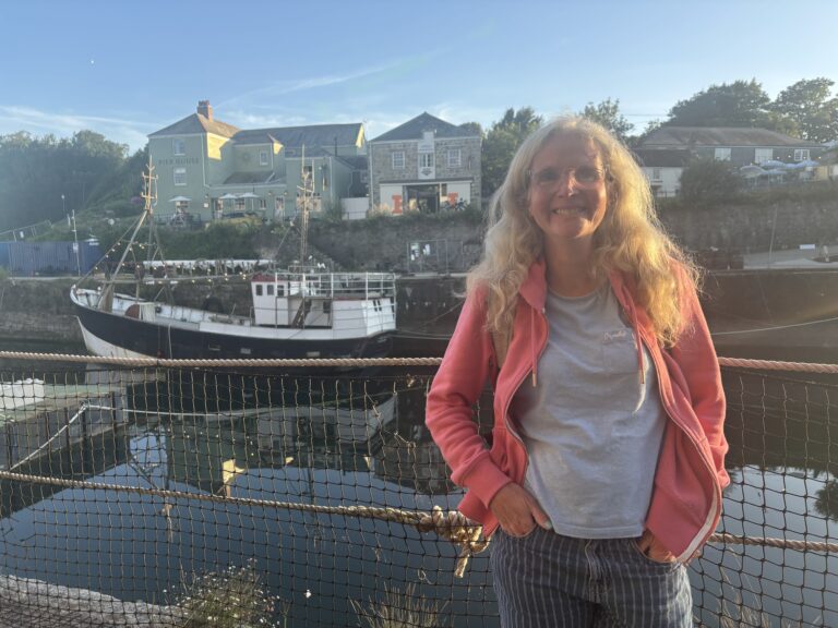 Charlestown harbour, Charlestown, Cornwall, A smiling woman in a pink hoodie and glasses stands by a harbour with a boat and buildings in the background
