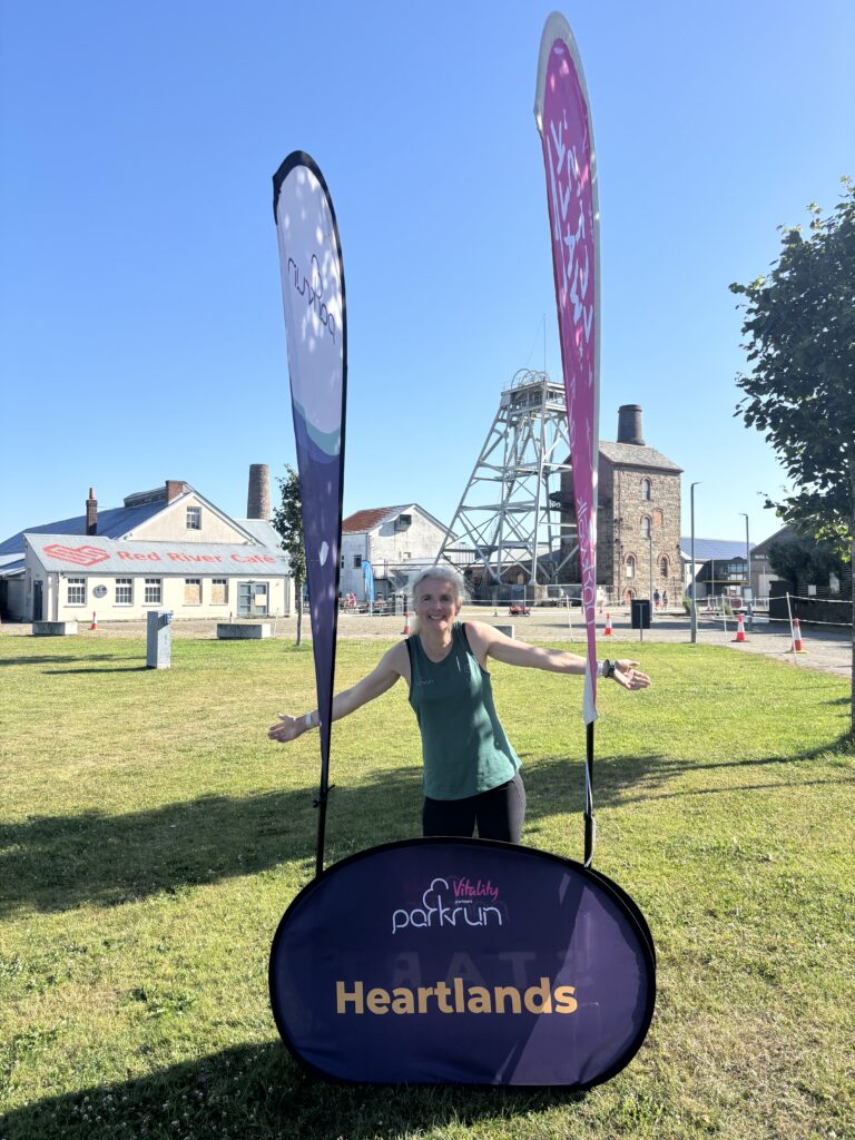 Heartlands parkrun, Parkrun, Cornwall, Redruth, Parkrun tourism, A female runner in a green vest stands behind a big purple sign which says Heartlands with buildings and a blue sky in the background