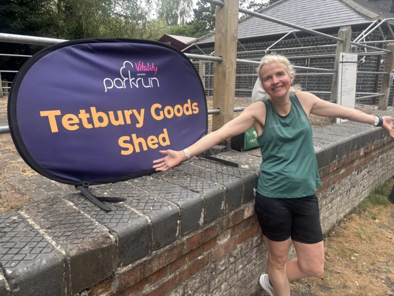 Tetbury Goods Shed parkrun, Parkrun, Parkrun tourism, A female runner in a green vest stands next to a big purple sign with Tetbury Goods Shed in yellow