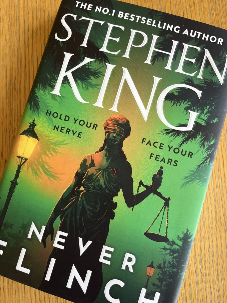Never Flinch by Stephen King, Never Flinch, Stephen King, Book review, A green book with a justice statue on the front called Never Flinch by Stephen King sits on a wooden table