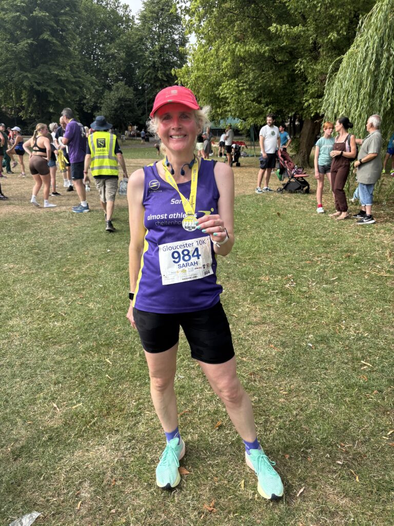 Gloucester 10k, Gloucester 10k medal, Running, Runner, A female runner wearing a purple vest and pink hat and holding a medal stands on the grass in front of other runners