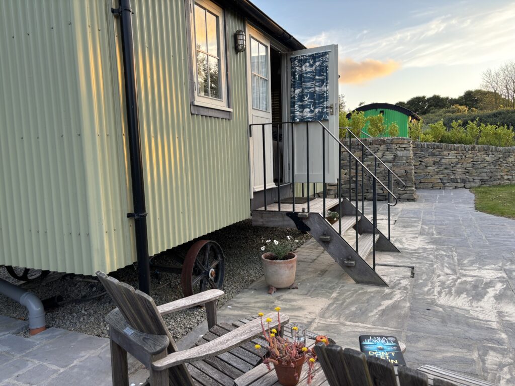 Rick Stein's shepherd's hut, Shepherd's hut, A light green shepherd's hut with the door open with wooden seating in the foreground and another shepherd's hut in the distance just before sunset