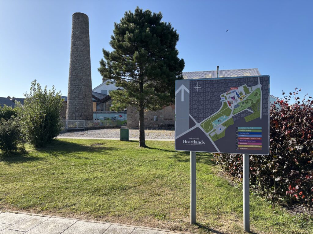 Heartlands parkrun, Parkrun, Cornwall, Redruth, A map on a signpost which says Heartlands in front of trees and hedges and a tall brick chimney against a blue sky