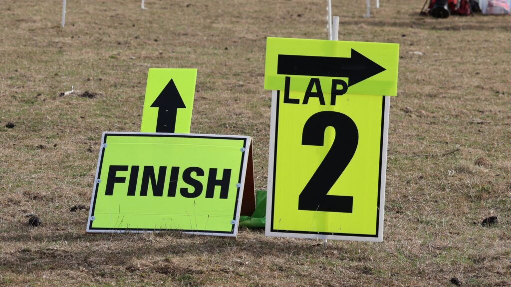 Thoulstone parkrun, Parkrun tourism, Two yellow signs sit next to each other on dead dry grass - one is an arrow and finish and the other is an arrow and lap 2