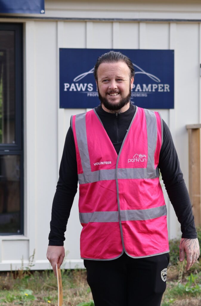 Thoulstone parkrun, Parkrun tourism, First timers' briefing, A smiling man with very dark hair and a dark beard wearing a pink bib saying volunteer over black clothing