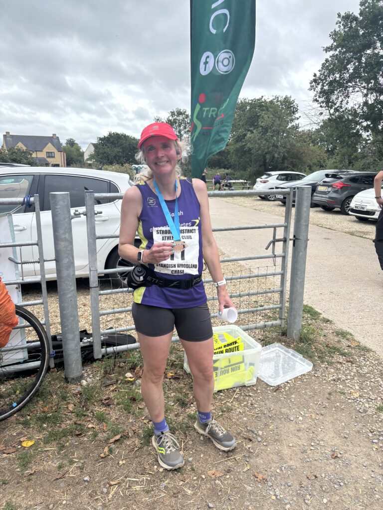 Standish Woodland Chase, Running, Runner, A female runner in a purple vest and pink hate wearing a small wooden medal standing in front of a metal gate a green flag and some cars