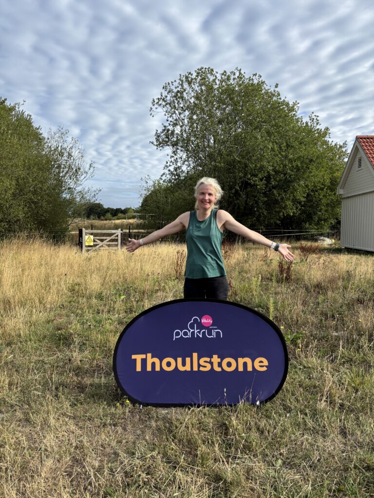 Thoulstone parkrun, Parkrun tourism, A smiling female runner in a green vest stands behind a purple sign saying Thoulstone in yellow lettering on long yellow grass with trees in the background