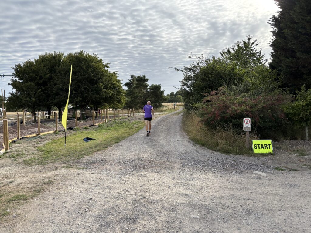 Thoulstone parkrun, Parkrun start line, Parkrun tourism, A small yellow start sign sits in front of a hedge next to a gravel path amongst trees and hedges with a yellow flag opposite and a single male runner in a purple top running away from the photographer