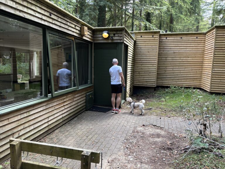 Center Parcs, Dog friendly lodge, A low wooden building surrounded by trees with a man in a white Tshirt and a small cockapoo dog about to go in the door