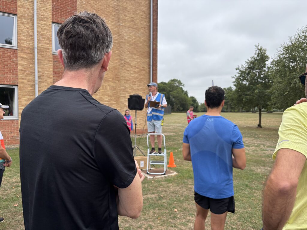 Fire Service College parkrun, Parkrun tourism, Run director's briefing, Back view of three men with a man wearing a blue bib standing on a small ladder facing the crowd in front of a building and some trees