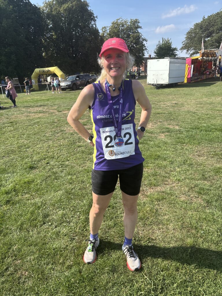 Pershore Plum 10k, Running, Runner, A smiling female runner in a purple vet and pink cap stands on the grass wearing a medal with a purple ribbon