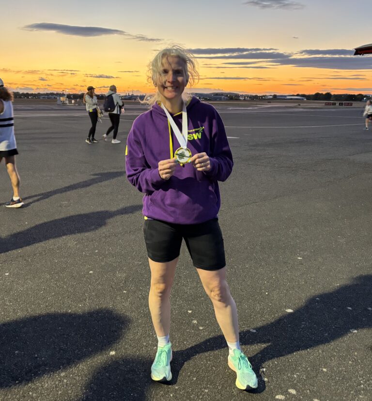 Meteor Mile, Running, Runner, Sunset, Gloucestershire Airport, A smiling woman in shorts and a purple hoodie wearing a medal stands on the runway of an airport at sunset