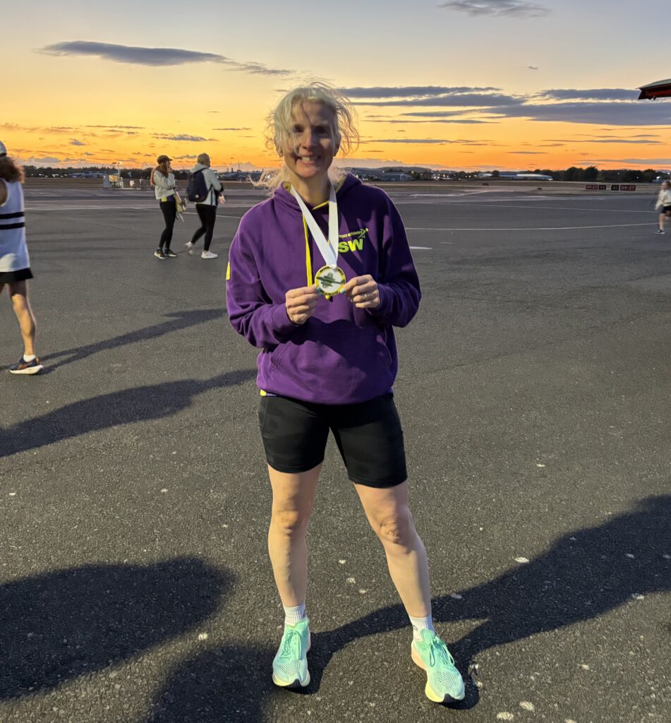 Meteor Mile, Running, Runner, Sunset, Gloucestershire Airport, A smiling woman in shorts and a purple hoodie wearing a medal stands on the runway of an airport at sunset