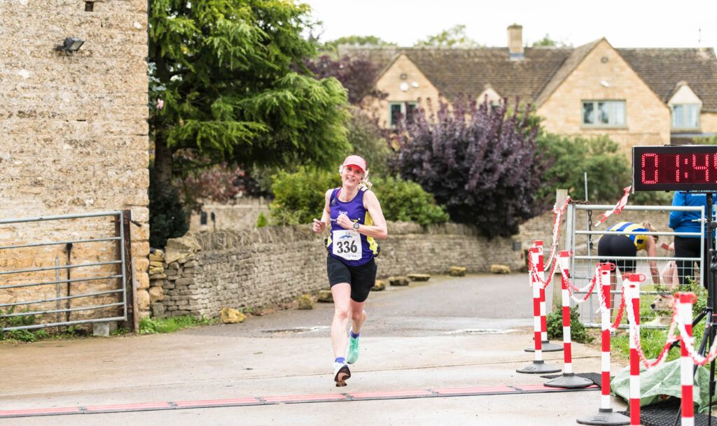 Bourton Hilly Half, Bourton half marathon, Finish line, A female runner in a purple vest and pink cap crosses a finish line with Cotswold stone cottages in the background and a big digital clock just visible