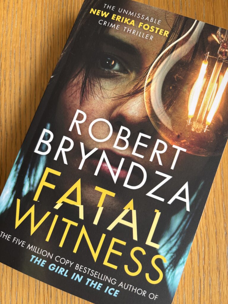Fatal Witness by Robert Bryndza, Fatal Witness, Robert Bryndza, Book review, A book called Fatal Witness by Robert Bryndza with a woman's face and a lightbulb on the cover sits on a wooden table