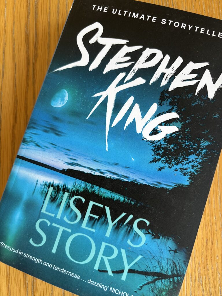 Lisey's Story by Stephen King, Lisey's Story, Stephen King, A blue book with water and a dark sky on the cover called Lisey's Story by Stephen King sits on a wooden table