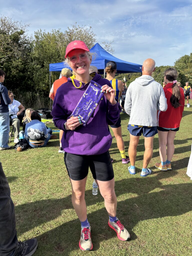 Cranham Boundary Chase, Running, Runner, A smiling woman in a purple hoodie and black shorts stands on grass in front of crowds of people and holds a large bar of chocolate wrapped in purple