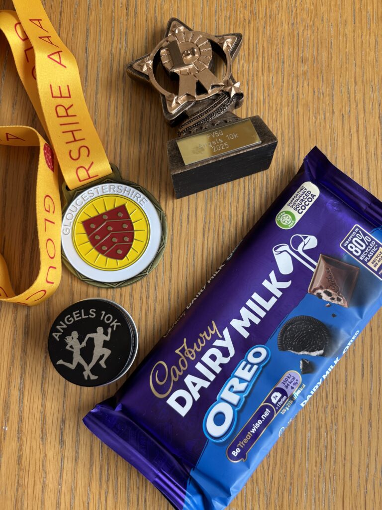 Angels 10k, Angels 10k prizes, Medal, Trophy, A bar of chocolate and a small trophy in the shape of a star and a medal with a yellow ribbon and a small black tin which says Angels 10k sit on a wooden table