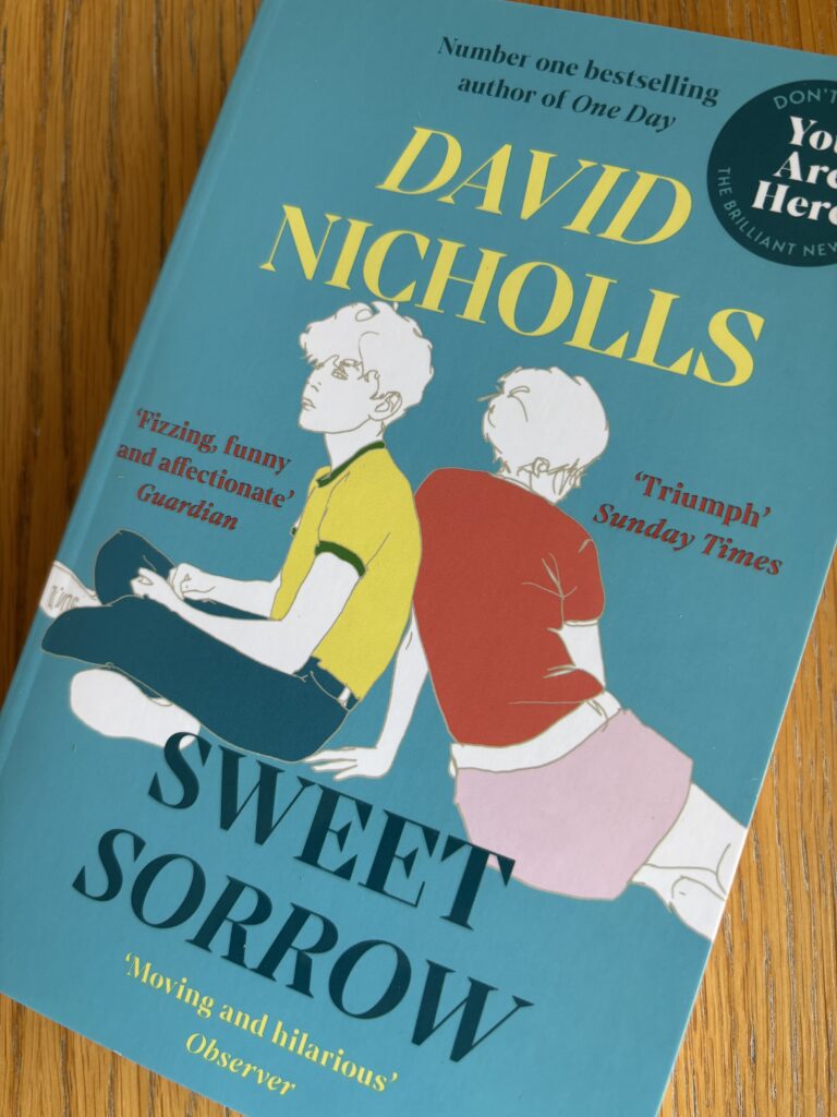 Sweet Sorrow by David Nicholls, Sweet Sorrow, David Nicholls, Book review, A blue book called Sweet Sorrow by David Nicholls with a drawing of two young people sitting down on the cover sits on a wooden table