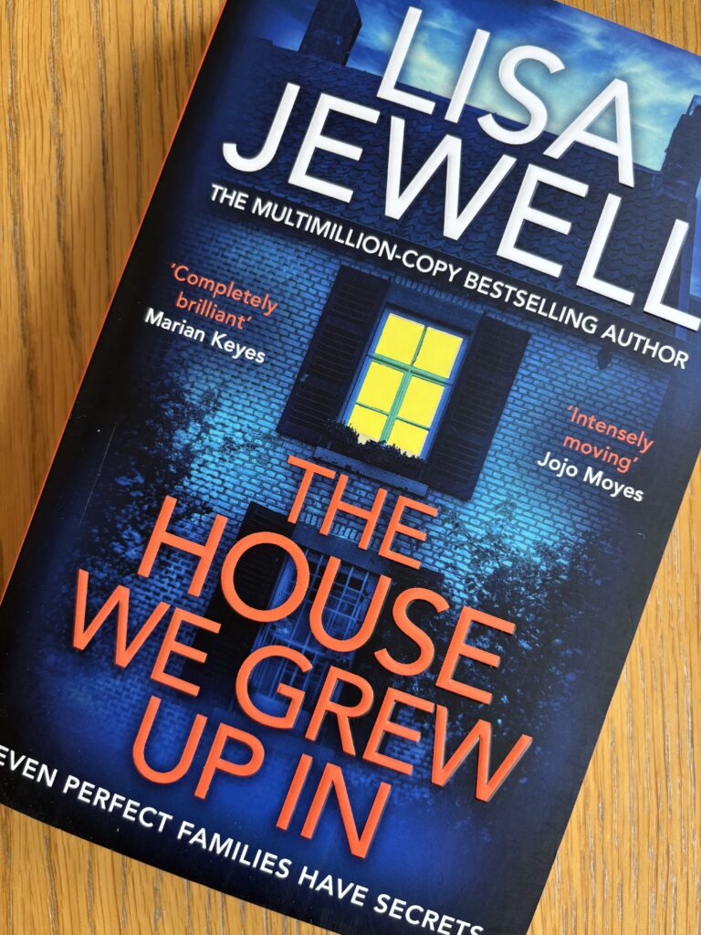 The House We Grew Up In by Lisa Jewell, The House We Grew Up In, Lisa Jewell, Book review, A blue book showing a house in darkness with a lit up window called The House We Grew Up In by Lisa Jewell sits on a wooden table
