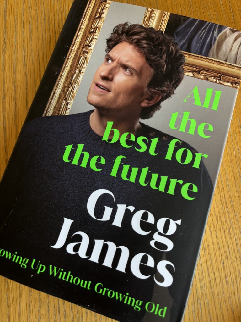 All the best for the future by Greg James, All the best for the future, Greg James, A book with a head and shoulders photo of a 30-something man with brown hair called All the best for the future by Greg James sits on a wooden table
