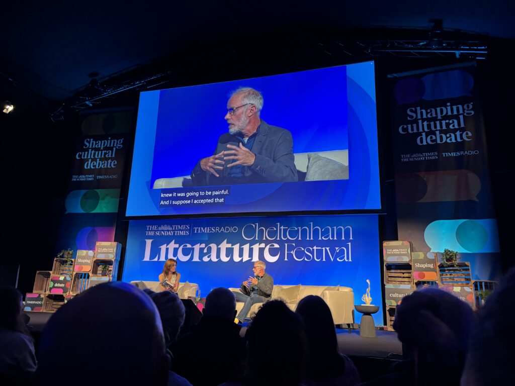 Paul Sinton-Hewitt, One Small Step, Cheltenham Literature Festival, A large blue screens shows a thin man with grey hair speaking to an audience above a sign that says Cheltenham Literature Festival and a stage where he is just visible on a sofa talking to a woman next to him