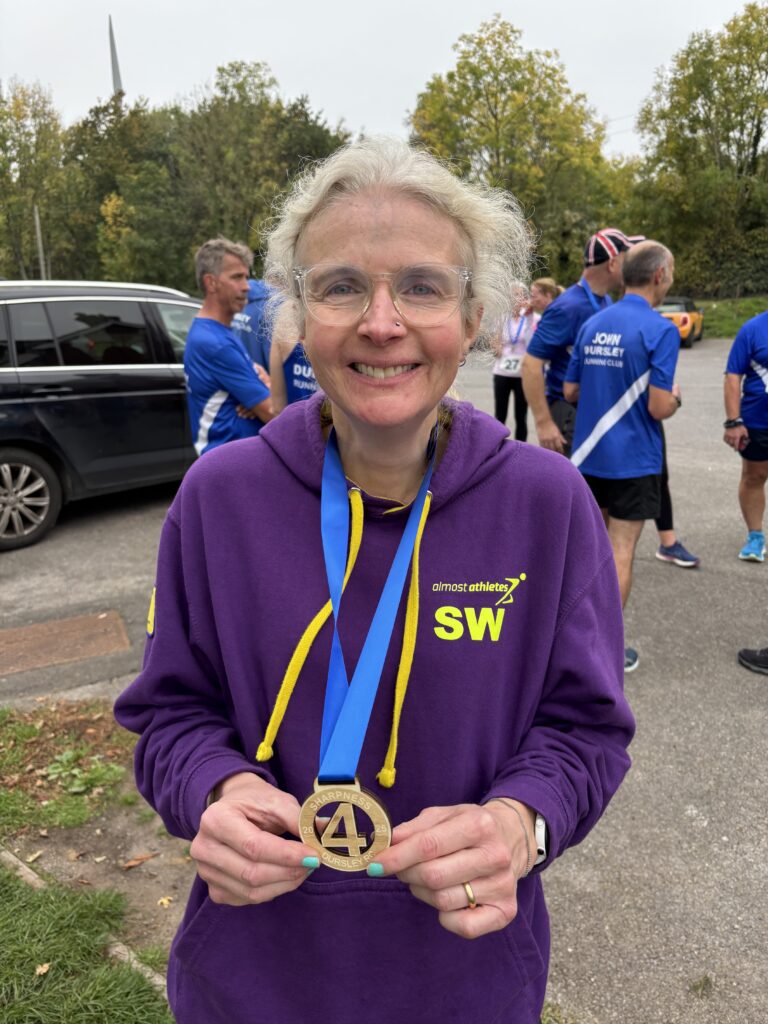 Sharpness 4, Sharpness 4 medal, Running, Runner, A female runner wearing glasses and a purple hoodie with a wooden medal round her neck and other runners in the background