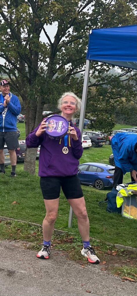 Sharpness 4, Running, Runner, A smiling female runner in a purple hoodie holds a purple tub of Heroes chocolates