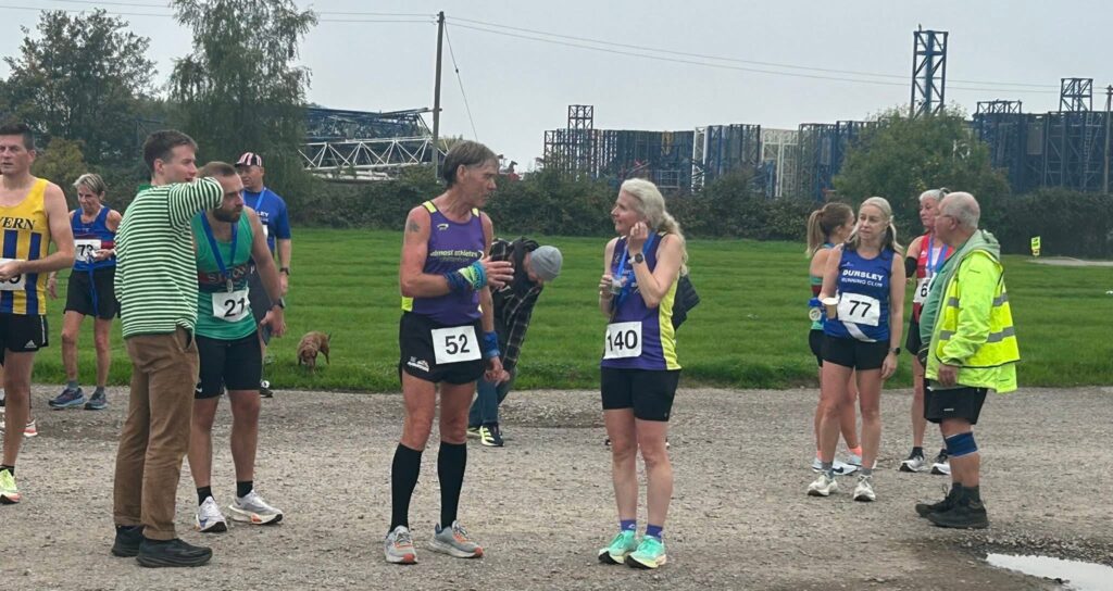 Sharpness 4, Sharpness Docks, Running, Runners, A group of runners stands on a gravel path in front of a patch of grass and industrial structures against a grey sky
