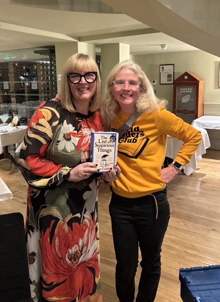 Cheltenham Literature Festival, Jennie Godfrey, The List of Suspicious Things, , Two middle aged women in glasses one wearing a floral dress and the other wearing a yellow jumper and black jeans stand in a large room holding a book called The List of Suspicious Things