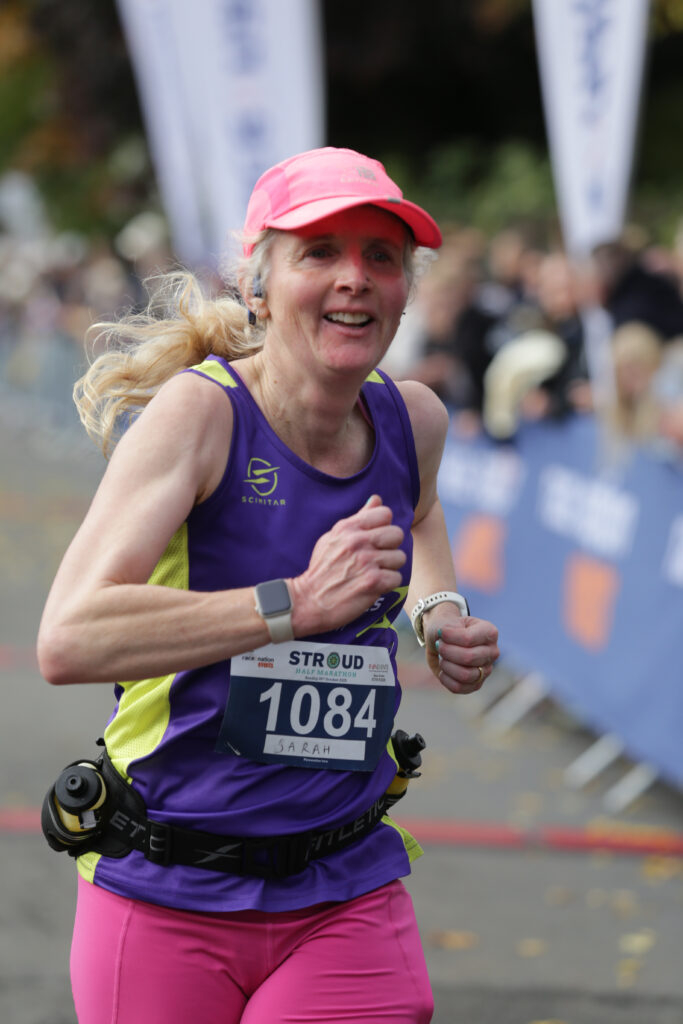 Stroud half marathon 2025, Stroud half, Running, Runner, Close-up photo of a female runner in a pink cap and pink shorts and a purple vest wearing a race number that says Stroud with flags and crowds visible in the background