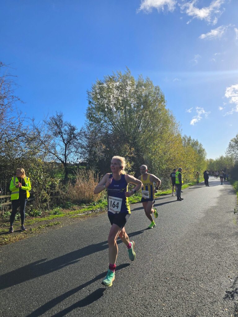 Guy Fawkes 5, Tewkesbury, Running, Runners, A female runner in a purple vest runs ahead of a male runner in a yellow vest along a road with trees and hedges in the background and a blue sky with other people in the background