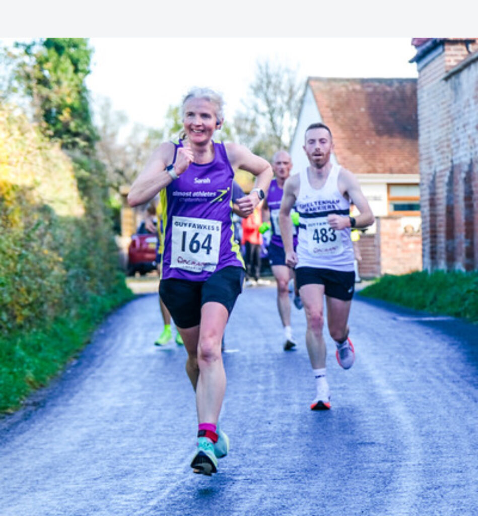 Guy Fawkes 5, Tewkesbury, Running, Runners, A female runner in a purple vest runs ahead of a man in a white vest along a road with buildings and hedges on either side and other runners in the background