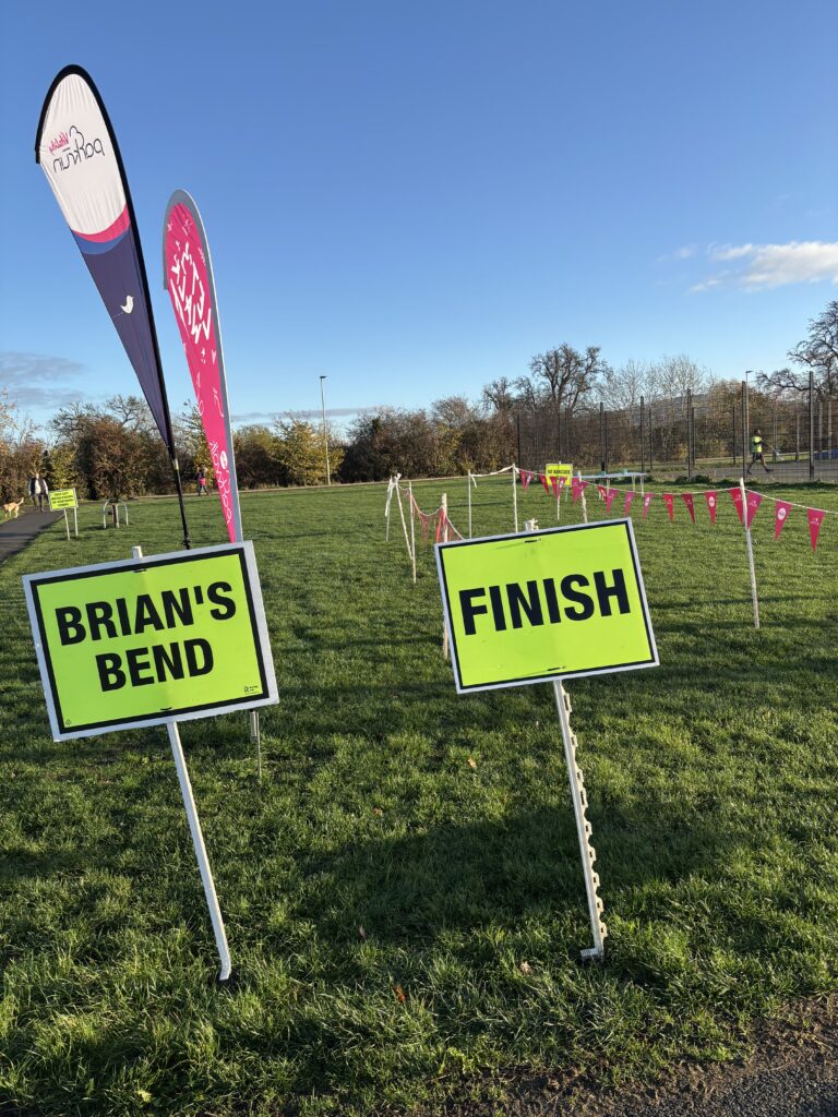 Kingsway parkrun, Parkrun, Parkrun tourism, Kingsway parkrun finish line, A row of bunting and two tall flags with a yellow sign saying finish on the grass against a blue sky with bare trees in the distance