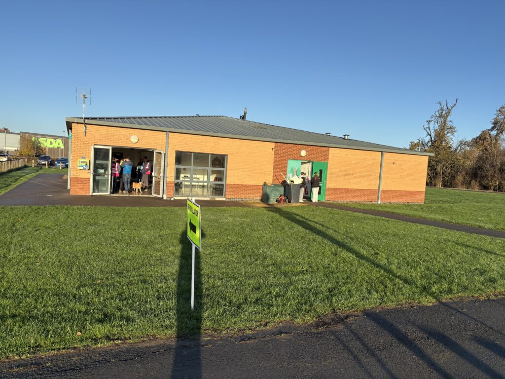 Kingsway parkrun, Parkrun, Parkrun tourism, A low brick building with open doors and people inside it is surrounded by grass against a blue sky with bare trees in the background