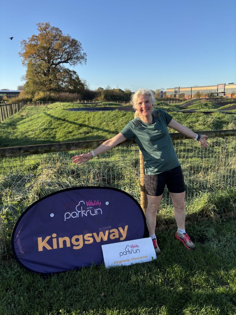 Kingsway parkrun, Parkrun, Parkrun tourism, A female runner in a green top stands on the grass next to a big purple sign which says Kingsway in yellow writing against a backdrop of grass and a big tree with brown leaves and a blue sky