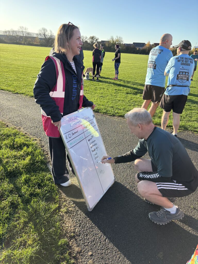 Kingsway parkrun, Parkrun, Parkrun tourism, A woman wearing pink hi-viz holds a big white board and a man crouches down to write on it and there are other people in the background standing on the grass against a blue sky