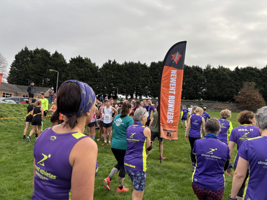 Newent 9, Running, Runners, A sports field with trees in the background and a grey sky and a tall black and orange sign which says Newent Runners with lots of runners