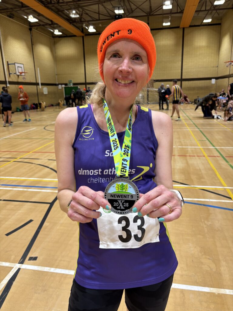 Newent 9, Newent 9 medal, Running, Runner, A female runner in a purple vest and an orange hat saying Newent 9 holds her medal standing inside a large sports hall