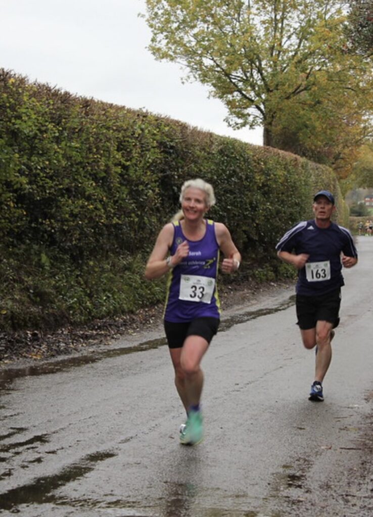 Newent 9, Running, Runners, A females runner in a purple vest runs in front of a male runner in dark running clothes along a very wet country lane with a tall hedge alongside it and a very autumnal tree in the background