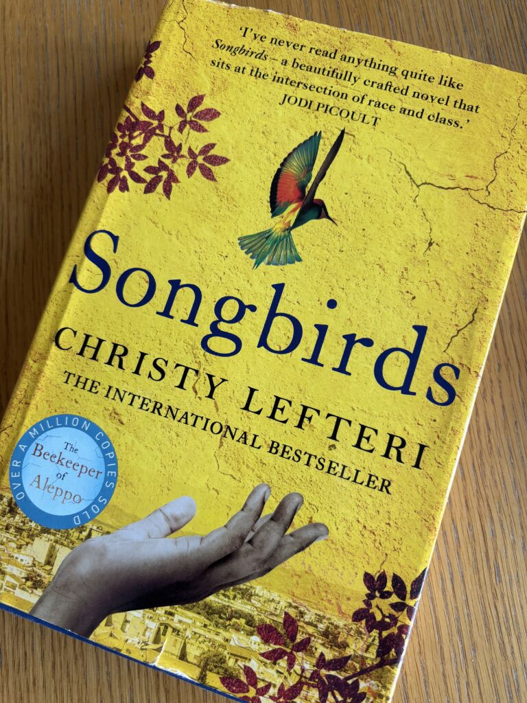 Songbirds by Christy Lefteri, Songbirds, Christy Lefteri, Book review, A yellow book with a hand and a colourful bird on the cover called Songbirds by Christy Lefteri sits on a wooden table
