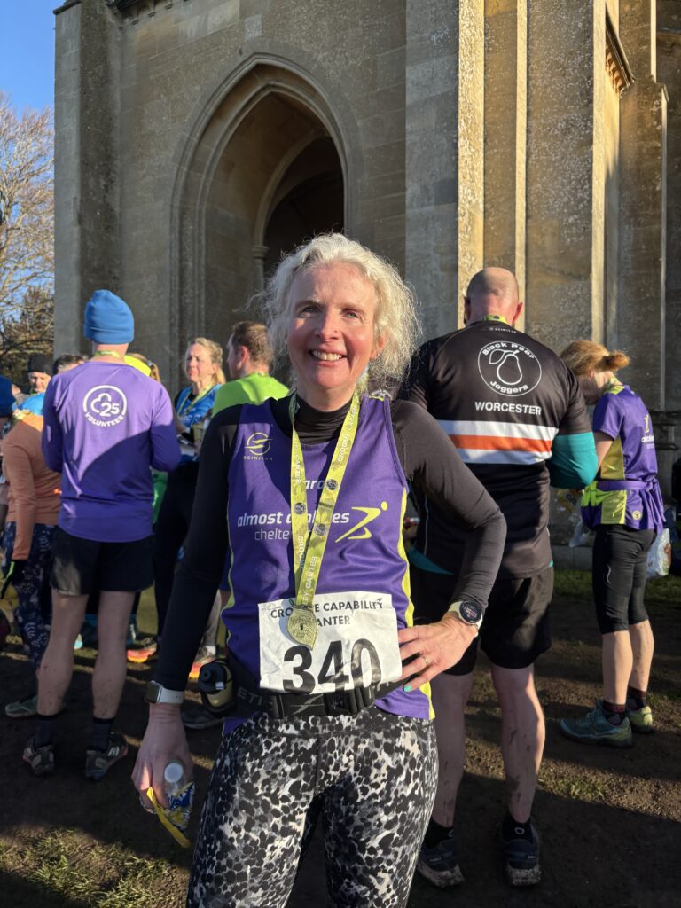 Croome Capability Canter, Runner, Runner, Croome Capability Canter medal, A smiling female runner wearing a purple vest over a black long sleeved top wears a medal in front of a crowd of runners outside a church