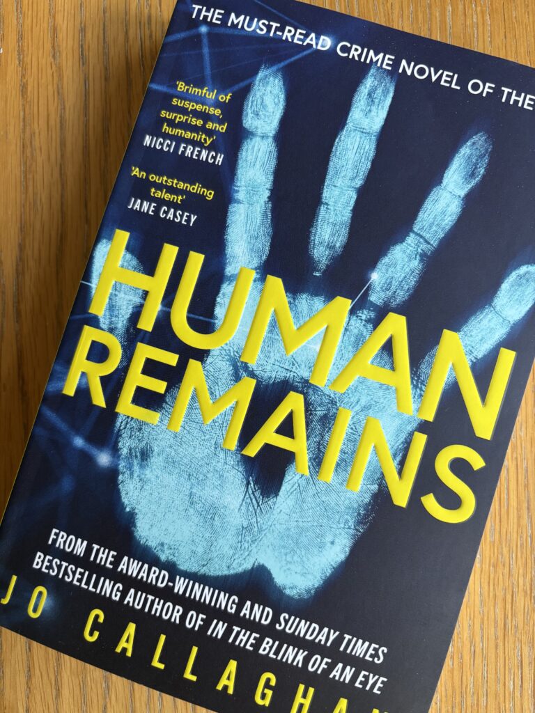 Human Remains by Jo Callaghan, Human Remains, Jo Callaghan, Book review, A black book Human Remains by Jo Callaghan with a big blue handprint on the cover sits on a wooden table