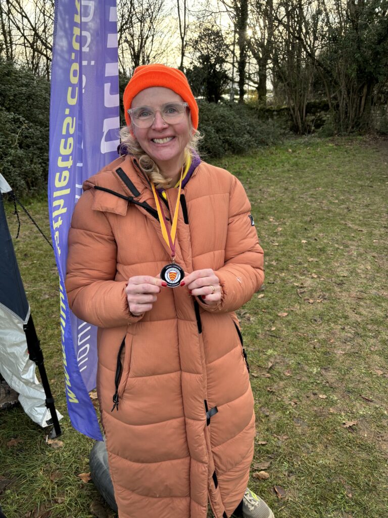 Cross country, Bath cross country, Running, Runner, A smiling woman in a padded coat, glasses and an orange hate wearing a medal standing on the grass in front of a tall purple flag